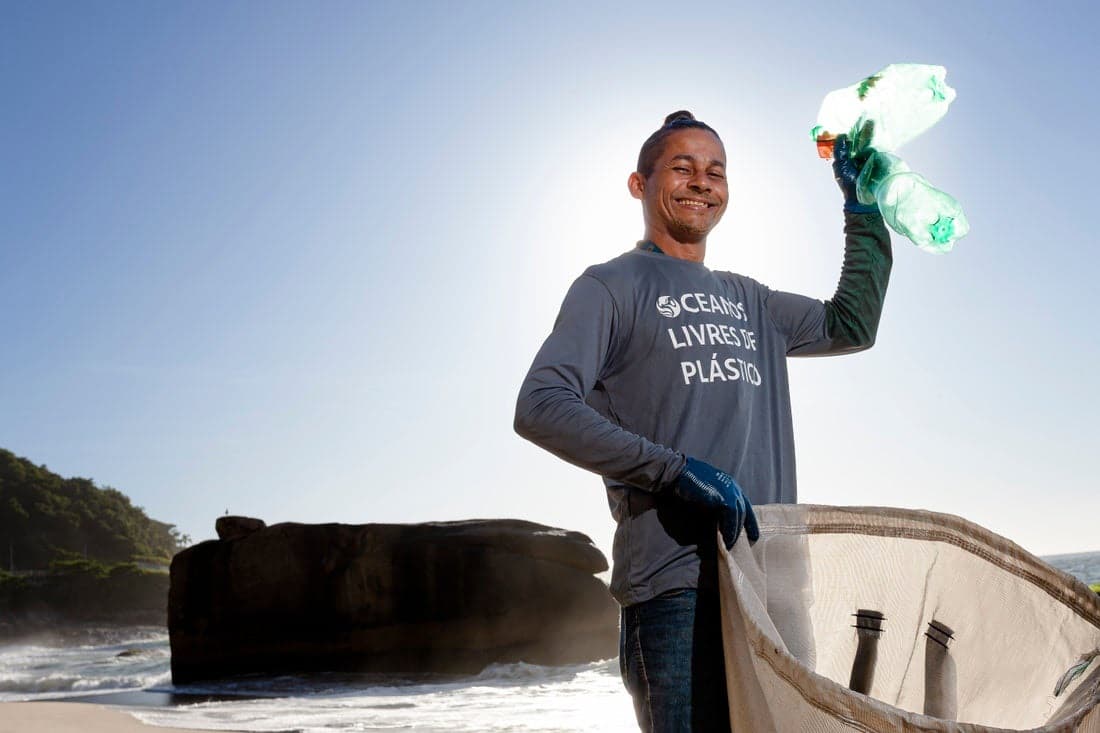 a smiling man contributing to environmental conservation by collecting plastic waste. He holds up a clear plastic bottle to the light, showcasing his find, while holding a mesh bag likely for gathering more litter.